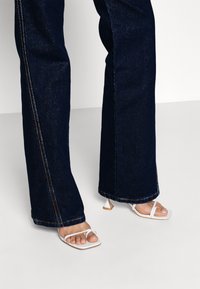 Jean flare taille haute en denim bleu foncé avec des coutures orange contrastantes, associés à des sandales à talons blancs avec un design à bout carré.