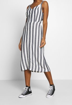 Gestreepte midi-jurk in marineblauw en wit, met dunne banden en een knoopsluiting aan de voorkant. Gecombineerd met zwarte hoge sneakers.