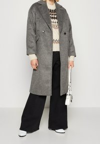 Persona con un abrigo largo gris abotonado, suéter crema con estampado, pantalones anchos negros, zapatos blancos y sosteniendo un pequeño bolso blanco.