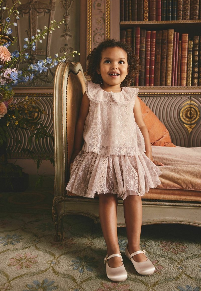 Mladé dievča v ružových čipkovaných šatách a saténových topánkach stojí usmievajúc sa vedľa ozdobnej vintage postele s knihami a kvetmi v pozadí.