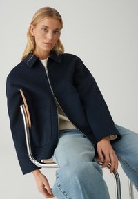 Giacca di lana blu navy con chiusura frontale a zip, colletto a punta e grandi tasche frontali. Indossata sopra una maglia color crema con jeans azzurri.