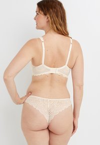 Set di lingerie in pizzo color beige chiaro; presenta bordi ondulati, spalline regolabili e un perizoma abbinato con dettagli in pizzo intricati.