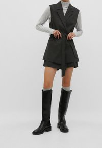 Blazer grigio a righe sottili senza maniche con cintura, sovrapposto a un dolcevita grigio, abbinato a stivali neri fino al ginocchio e calzini grigi.