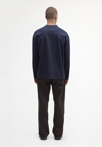 Granatowa koszula z długim rękawem, okrągłym dekoltem i luźnym krojem, zestawiona z brązowymi spodniami o prostym kroju, widok z tyłu.