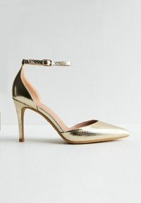 Zapato de tacón alto dorado con punta afilada y acabado texturizado de reptil. Presenta una correa en el tobillo con hebilla y un delgado tacón de aguja.