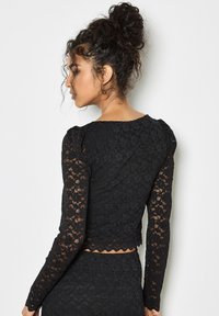 Svart spetstopp med långa ärmar som har en scalloped nederkant och texturerat blommönster, med puffiga axlar och en figurnära passform.