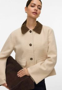 Veste beige en m élange de laine avec un col en velours côtelé marron, dotée de boutons arrondis et de grandes poches plaquées ; le mannequin tient une pochette marron foncé.