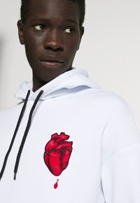 White hoodie with a red embroidered heart design on the left chest. Features black drawstrings and smooth cotton material.