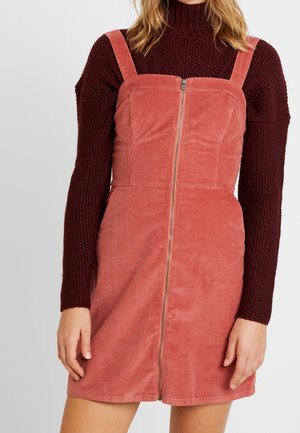Woman wearing a sleeveless dusty pink corduroy dress with front zipper over a dark maroon knitted long-sleeve turtleneck sweater.