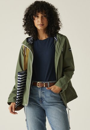 Vrouw met krullend haar, een groen jack, een marineblauw shirt, een blauwe spijkerbroek en een bruine riem, met een marineblauw-wit gestreepte tas over haar schouder.