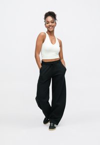 Juodos sportinės kelnės su raišteliu liemens srityje, derinamos su aptemptu balto spalvos marškinėlių be rankovių viršumi. Modelis stovi atsipalaidavęs, rankos kišenėse. Juodi sportiniai batai su baltais dryžiais.