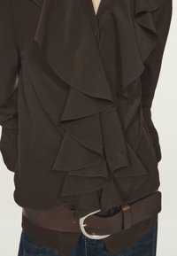 Brązowa bluzka z falbankami, z głębokim dekoltem w kształcie litery V, długimi rękawami oraz teksturowanym paskiem z srebrną klamrą noszonym w talii.