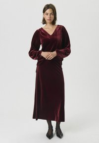 Vestido largo de terciopelo burdeos de manga larga con escote en V, cintura ajustada y falda acampanada. Cuenta con mangas abullonadas y zapatos negros de punta afilada.