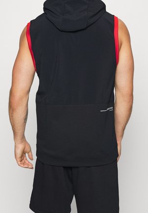 Sudadera sin mangas negra con detalles en rojo, presenta una textura suave, capucha y ajuste holgado. Vista trasera muestra un acabado liso y una marca mínima.