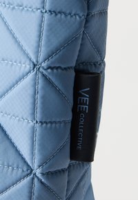 Ανοιχτό μπλε υφασμάτινο με ραφές σε σχήμα διαμαντιού και μια μαύρη ετικέτα στη πλευρά με μπλε γράμματα που αναγράφουν "VEE COLLECTIVE".