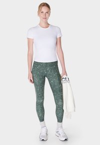 Vrouw in een witte aansluitende t-shirt, groene gespikkelde legging, witte sneakers en sokken, die een opgevouwen witte sweatshirt naast zich houdt.