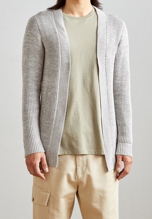 Cardigan côtelé gris clair avec une coupe ouverte, des manches longues et un tissu texturé, porté sur un t-shirt vert sauge et un pantalon beige.