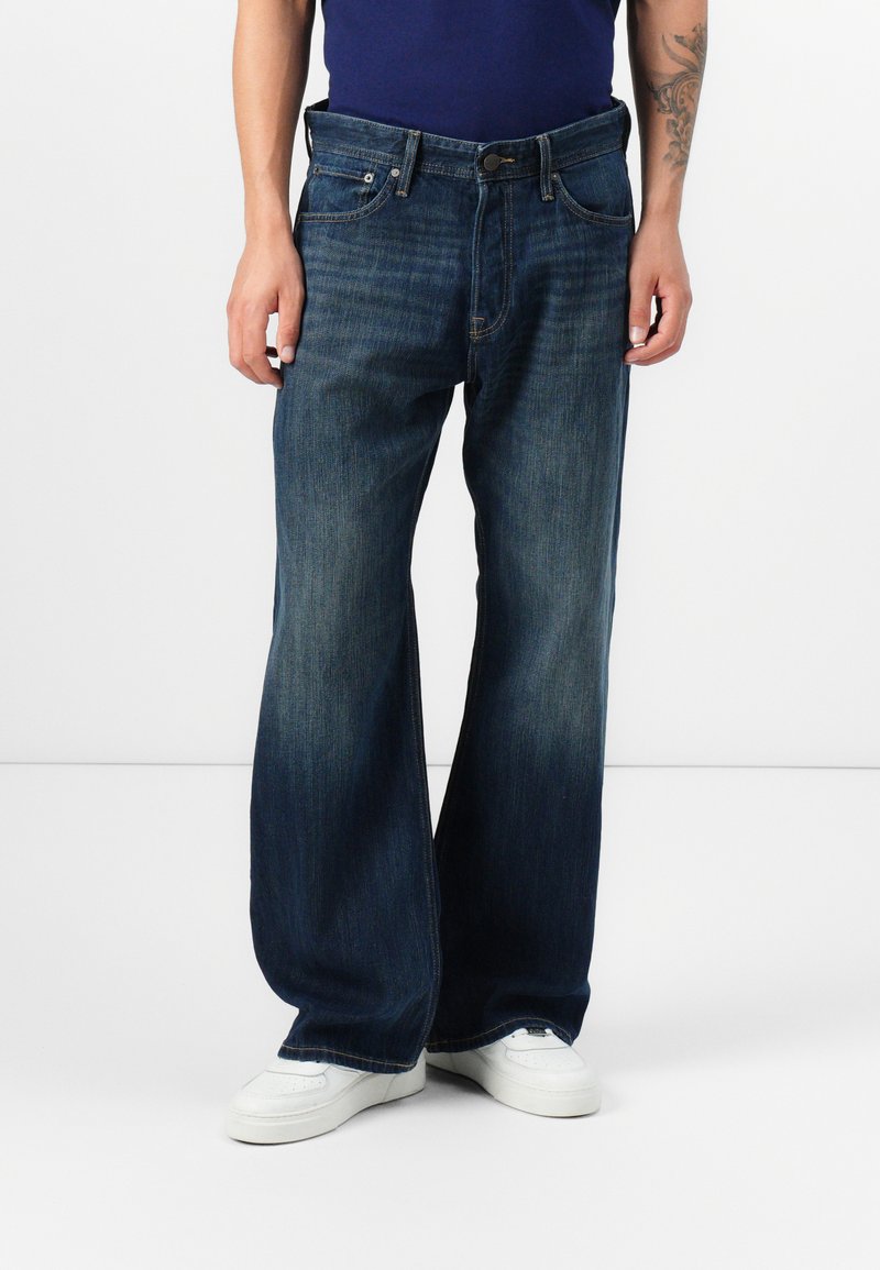 Jean bleu foncé à coupe large, fabriqué en denim. Comprend cinq poches et une fermeture à bouton. Porté avec des baskets blanches.