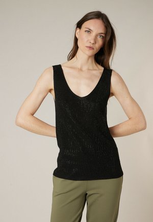 Femme aux longs cheveux bruns portant un haut en tricot noir sans manches avec un col en V et un pantalon vert olive, debout devant un fond uni.