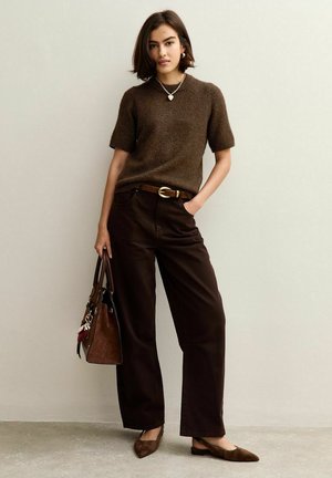Bruine gebreide trui met korte mouwen, brede bruine broek, gouden riem, platte bruine schoenen en een bruine handtas met accenten. Casual, samenhangende look.