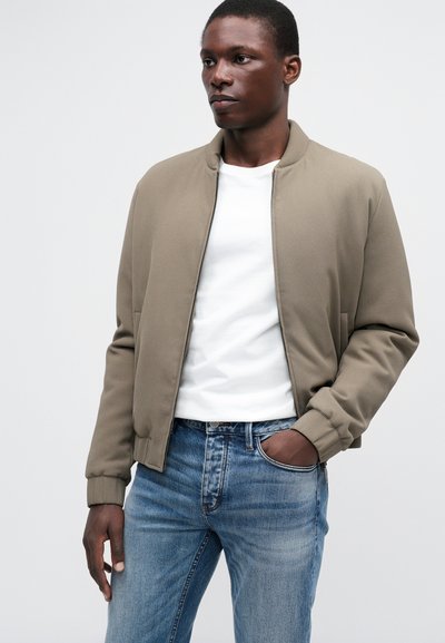 Kreminė bomber tipo striukė su tekstūruotu paviršiumi, raukšlėtomis rankovėmis ir golfo apykakle. Dėvima virš baltos marškinėlių ir derinama su mėlynomis džinsomis.