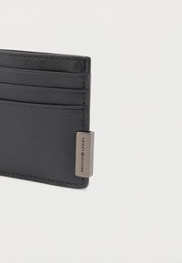 Portafoglio in pelle nera con quattro slot per carte e un'etichetta metallica incisa con il nome del marchio su sfondo bianco.