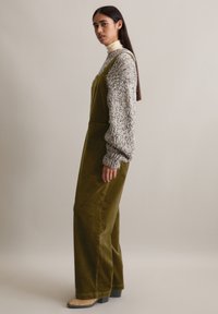 Olivengroene corduroy tuinbroek met wijde pijpen, gecombineerd met een grijze gebreide trui. Model draagt beige blokhak schoenen. Zachte, gestructureerde stoffen.