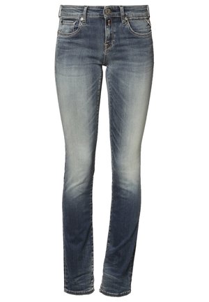 Femme portant un chemisier blanc à manches courtes, jean skinny bleu délavé, talons hauts noirs, tenant un chapeau noir dans une main.