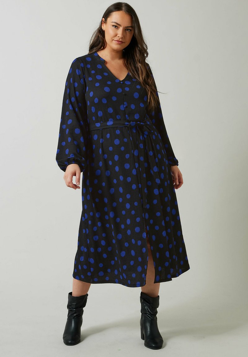 Musta mekko, jossa on suuria sinisiä ympyröitä, V-aukko, pitkät hihat, vyötärönauha ja polvimittainen pituus, yhdistettynä mustiin nilkkureihin.