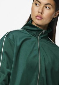 Grøn zip-jakke med høj krave, der har hvide sidelister og en glat, blank overflade. Sølv lynlåsdetalje synlig.