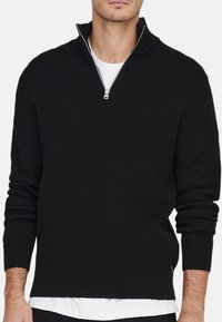 Sort striktrøje med halv-zip krave, ribbet tekstur og lange ærmer. Båret over en hvid t-shirt for ekstra stil.