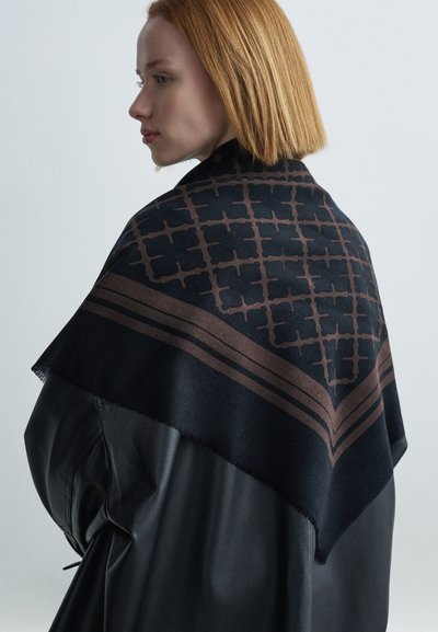 Sötét sál barna geometriai mintával, háromszög alakú díszítéssel és rojtos szegélyekkel, fekete bőr ruhadarabra viselve.