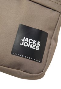 Pochette en textile marron avec une surface texturée. Présente une étiquette noire avec un texte blanc : "JACK & JONES ÉTABLI EN 1990." Design simple.