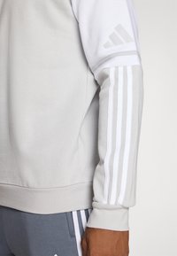 Sweat-shirt gris clair avec des accents blancs, arborant trois bandes blanches sur la manche et un design texturé et sportif.