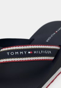 Navy flip-flop with a textured strap featuring red, white, and gold stripes. "TOMMY HILFIGER" printed on the strap in white text.