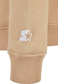 Manche de sweat-shirt beige avec un "S" brodé en blanc et un logo étoile au-dessus du bord-côte.