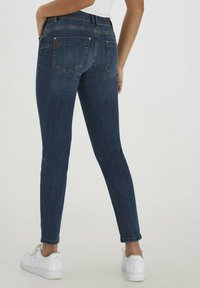 Lieknos tamsiai mėlynos džinsinės kelnės su vidutinio aukščio liemeniu, dviem užpakalinėmis kišenėmis ir subtiliais siuvinėjimo akcentais. Derinamos su baltais sportbačiais.