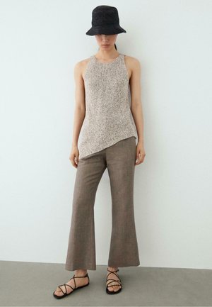 Kobieta w beżowym, bezrękawnym, dzianym topie z asymetrycznym dołem, brązowych szerokich spodniach, czarnym kapeluszu typu bucket i czarnych sandałach na paski, stojąca na tle białej ściany.