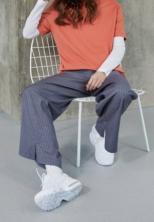 Koralowy t-shirt z krótkim rękawem nałożony na białą koszulkę z długim rękawem, zestawiony z szarymi, szerokimi spodniami w wąskie paski oraz białymi trampkami.