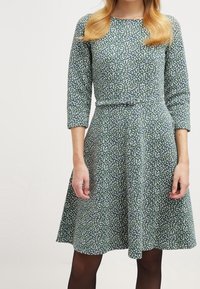 Vihreä kuvioitu mekko, jossa on kolmen neljänneksen hihat, korostettu vyötärö ja liehuva hame; siinä on pieniä valkoisia ja sinisiä kukkakuvioita.