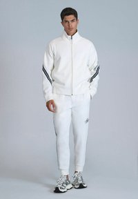 Weiße, mit Reißverschluss versehene Sportjacke mit schwarzen Streifenakzenten, kombiniert mit passenden weißen Jogginghosen mit Seitenstreifen und einem Logodetail.