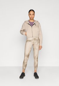 Μπεζ cropped φούτερ με μπροστινό φερμουάρ και λογότυπο, σε συνδυασμό με κάμελ κολάν με μοτίβο tie-dye. Μαύρα αθλητικά παπούτσια ολοκληρώνουν την εμφάνιση.