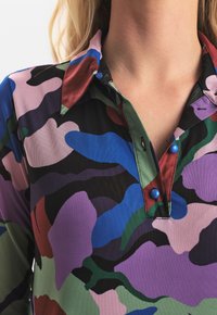 Färggad skjorta med krage som har ett abstrakt kamouflage-mönster i lila, blått, grönt och svart med två blå knappar vid halslinningen.