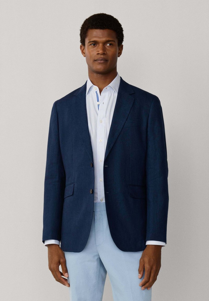 Mann trägt ein marineblaues Sakko, hellblau gestreiftes Hemd und hellblaue Hose, steht vor einem schlichten hellgrauen Hintergrund.