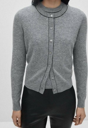 Nainen pukeutuneena istuvaan harmaaseen, mustilla reunoilla varustettuun napitettuun neuletakkiin ja mustiin housuihin, seisomassa yksinkertaista vaaleaa taustaa vasten.