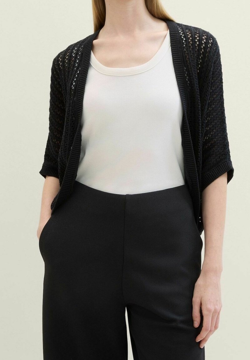 Cardigan nero lavorato a maglia con trama aperta, maniche corte, indossato sopra un top bianco aderente, abbinato a pantaloni neri a vita alta.