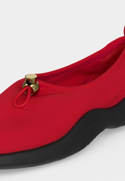 Κόκκινο παπούτσι slip-on με υφασμάτινο πάνω μέρος με υφή, μαύρη παχιά σόλα και χρυσή λεπτομέρεια toggle σε ελαστική θηλιά κοντά στο δάχτυλο.