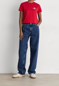 T-shirt rossa in cotone con un logo sul petto, abbinata a jeans in denim blu a vestibilità ampia. Indossata con sneakers di colore chiaro.