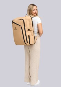 Mochila beige con acentos y logo negros, forma rectangular, correas acolchadas, gran compartimento principal, múltiples bolsillos con cremallera.