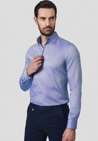 Jasnoniebieska koszula z długim rękawem, z delikatną fakturą, z kołnierzykiem w kształcie punktu, noszona z ciemnymi spodniami. Zawiera białe guziki.